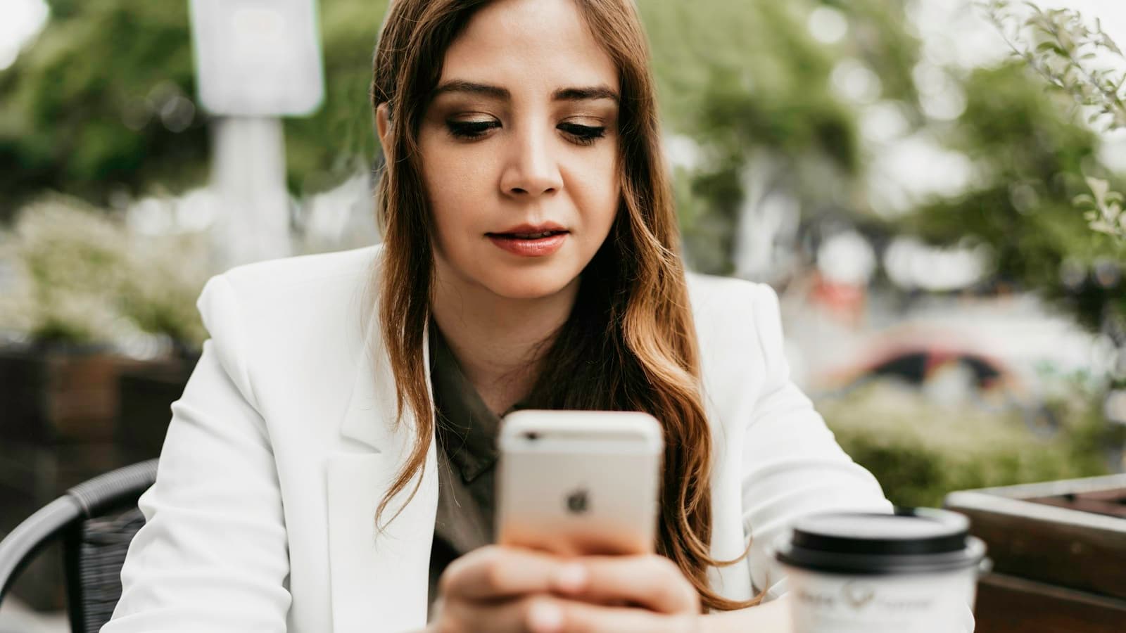 Mulher de blazer branco olhando o celular, com expressão séria, sentada num café